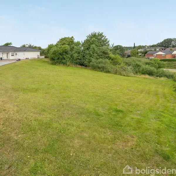 Græsplæne strækker sig mod en hvid bygning i baggrunden, omgivet af træer og buske, med flere huse synlige i det fjerne på en solrig dag.