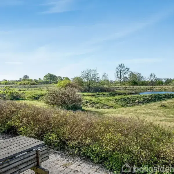 Et rødt hus med gråt tag står ved en sø. Omkringliggende græs og buske skaber et naturligt landskab under en klar, blå himmel.