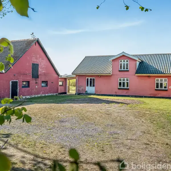 Rødt hus med tagvindue står stille i en gårdhave omkranset af græs og en blå himmel. En tilstødende rød bygning ses til venstre, omgivet af grønne træer.
