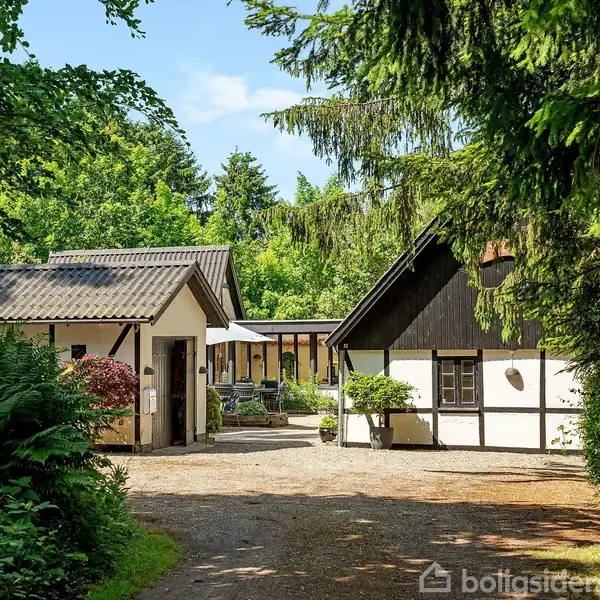 To huse i en skovrydning, omgivet af frodig vegetation. Den ene bygning har sort bindingsværk og hvid facade, den anden har skråt tag. Grusvej i forgrunden.