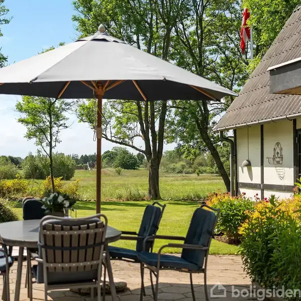 Parasol står over havebord med seks stole på terrasse. Omgivet af grønne træer og velplejet græsplæne, ligger det ved siden af et stråtækt hus med Dannebrog på taget.
