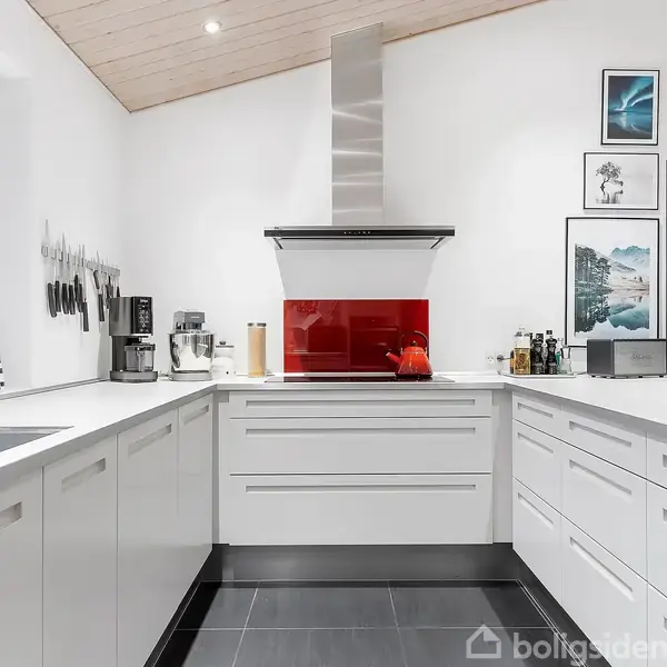 Køkken med hvide skabe omkring rødt komfur og emhætte; køkkenmaskiner og knive til venstre. Væggene har naturbilleder indrammet. Vindue til venstre slipper dagslys ind.