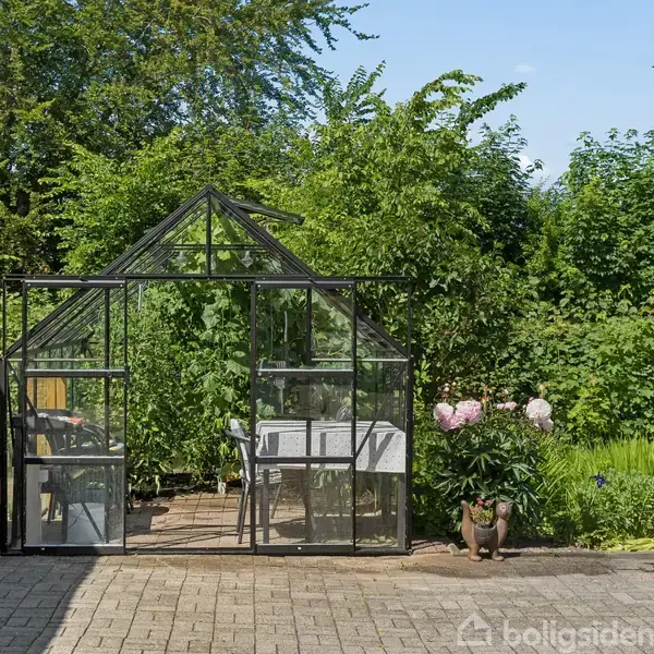 Et drivhus med glasvægge står på en flisebelagt terrasse. Indenfor ses en stol og et bord, omgivet af frodige grønne buske og blomster i en solrig have.