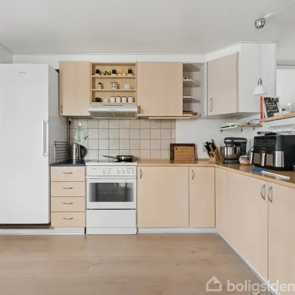 Køkken med lyse træskabe, kogeplade og ovn, placeret ved siden af høj køleskab. Køkkenbord med kaffemaskine og blender, flisebeklædt væg. Lys hænger fra loftet, og køkkenredskaber står ved bordet.