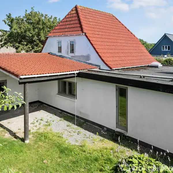 Hus med hvid facade og rødt tegltag har en terrasseoverdækning. Haven er græsbelagt med omkringliggende buske. Flere villaer og træer ses i nabolaget under en blå himmel.