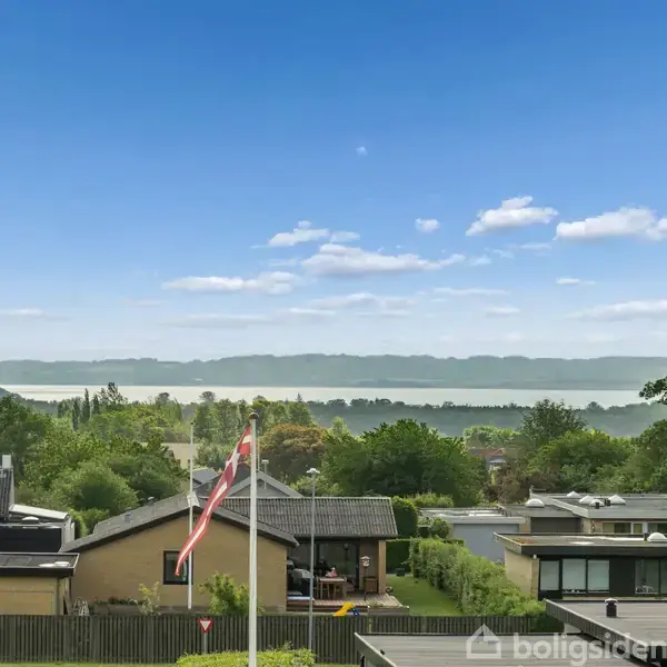 Hus tage med grønne haver ligger i forgrunden, mens en bred sø og skove kan ses i baggrunden under en blå himmel.