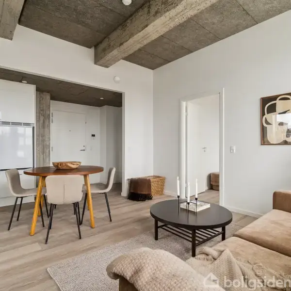 En moderne stue med en beige sofa og et rundt sort sofabord. I baggrunden står et rundt spisebord med fire stole i det åbne køkkenområde. En plante står i hjørnet.