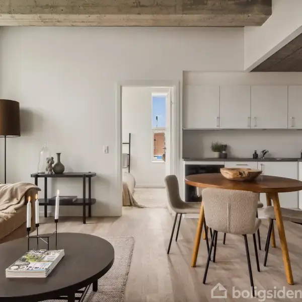 En moderne stue med en beige sofa til venstre, et rundt spisebord og stole i midten, og et køkken med hvide skabe i baggrunden. Flot træloft og gulv.