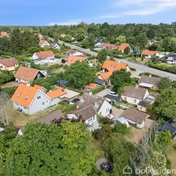 Et parcelhuskvarter med huse med røde tage spreder sig langs snoede veje og frodige haver. Området er omgivet af tæt bevoksning og spredte træer, der giver en grøn atmosfære.