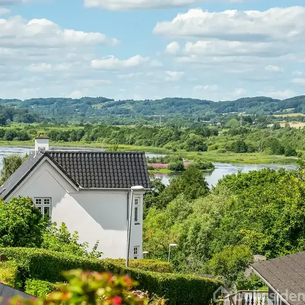 Et hvidt hus står stille omgivet af frodig grøn vegetation, med en flod og bakkede landskaber i baggrunden under en blå himmel med spredte skyer.