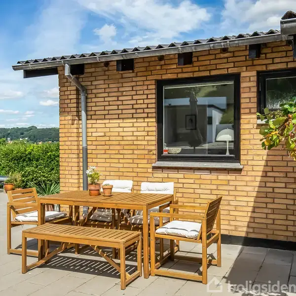 Et træbord med stole og en bænk står på en terrasse foran en gulstensmur. En plante hænger ved siden af et vindue. Landskab ses i baggrunden under en blå himmel.