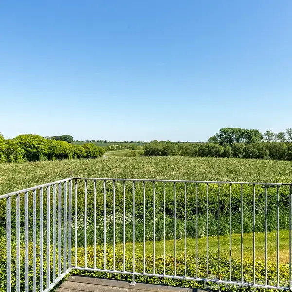 Et metalrækværk omgiver en balkon med udsigt over en grøn mark og horisonten, omgivet af træer under en klar blå himmel.