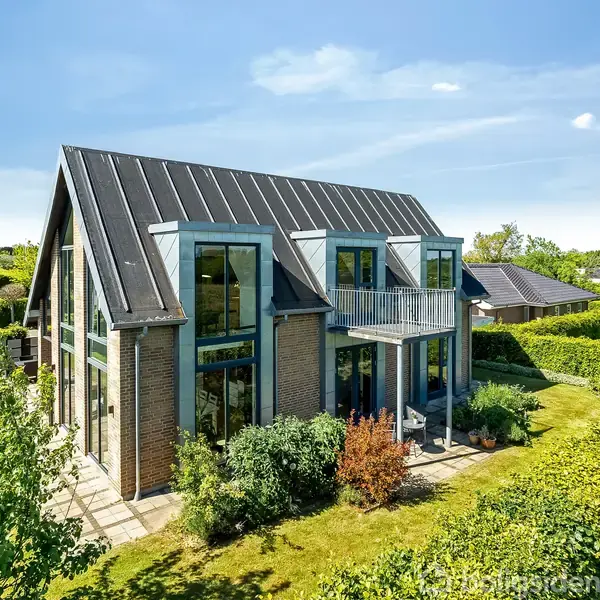 En moderne to-etagers villa med store glaspartier står i en grøn have omkranset af hække. Huset har en skrånende metaltag og balkon, beliggende i et beboelsesområde med andre huse.