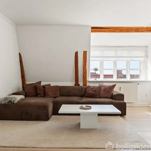Brun hjørnesofa med puder står på et lyst tæppe ved et lavt, hvidt sofabord. Store vinduer bagved giver naturligt lys, og træbjælker i loftet skaber en hyggelig atmosfære.