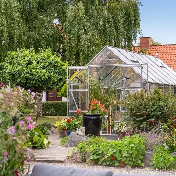 Drivhus omgivet af frodige planter og blomster, stående i en velplejet have med grønne træer og en murstensbygning med rødt tag i baggrunden.