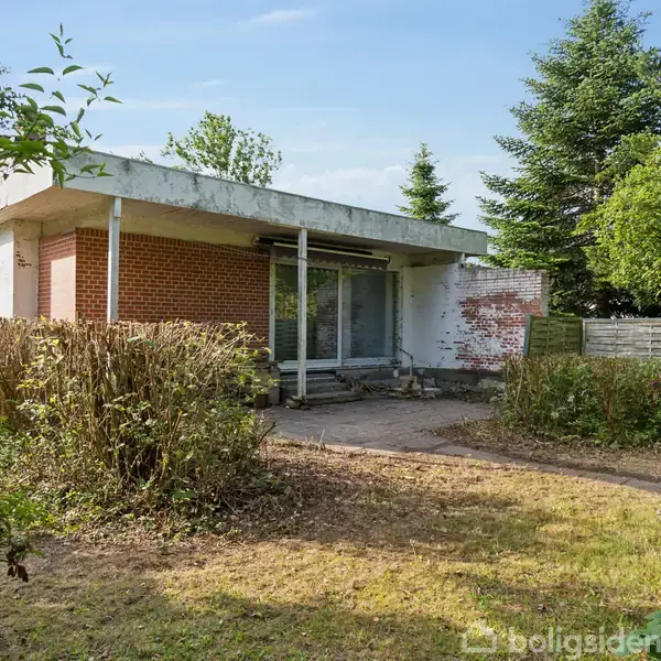 En enkel-etagers murstensbygning med fladt tag står midt i en have med buske og træer omkring sig. Bygningen har en terrasse foran store vinduer.