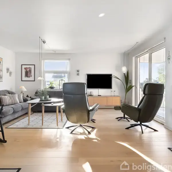 En stue med sorte lænestole og en grå sofa står oplyst af naturligt lys fra store vinduer. Et tv er placeret på et mediecenter, omgivet af lyse træmøbler.