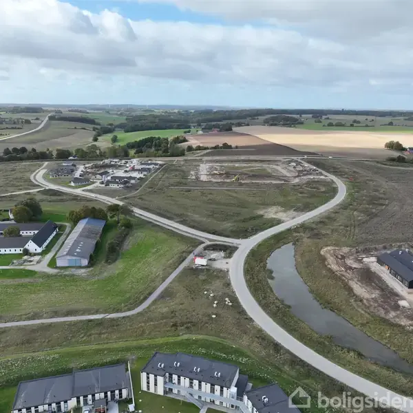 En snoet vej forbinder flere bygninger og et byggepladsområde i et landligt landskab med marker og træer under en overskyet himmel.