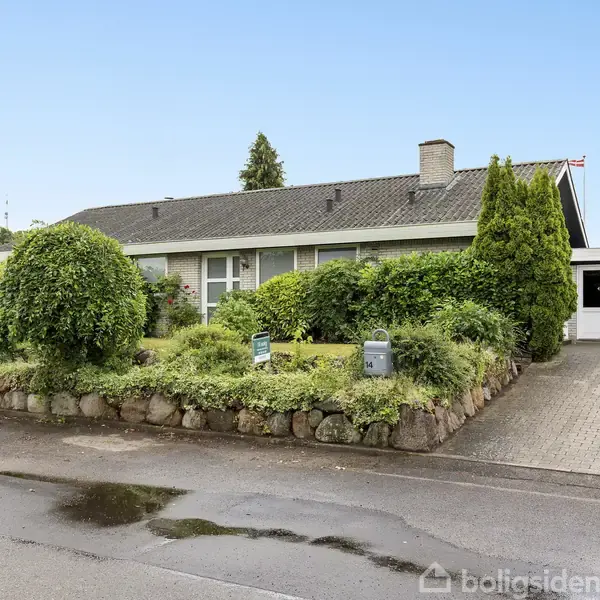 Et enfamiliehus med gråt tag, omgivet af velplejet have og buske, står ved en asfalteret indkørsel. Huset er placeret i et roligt kvarter.