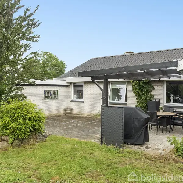 Et enfamiliehus med hvid murstensfacade har en terrasse i baghaven med et sort havesæt og grill. Haven er omkranset af grønne planter og en velplejet græsplæne.
