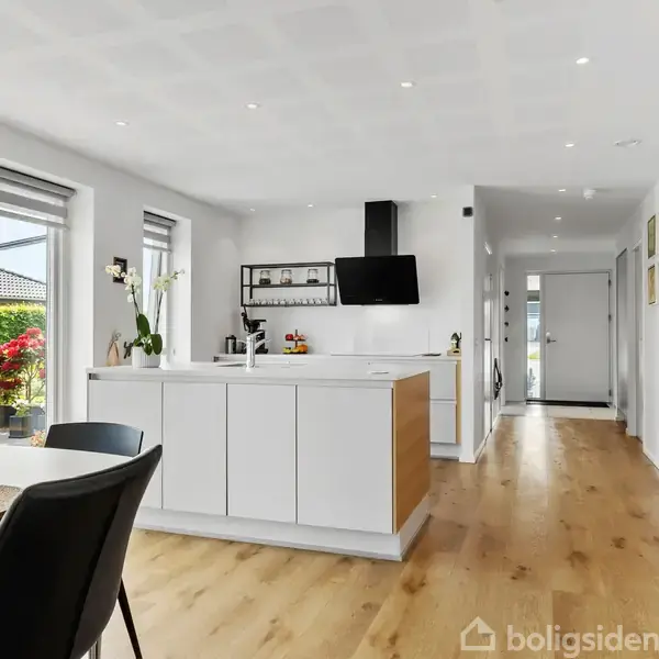 Et moderne køkken med en central ø og hvide skabe. Køkkenet er forbundet til en spisestue med bord og stole. Store vinduer giver udsigt til en have.