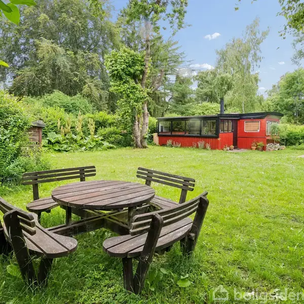 Rundt havebord med bænke står på en græsplæne. I baggrunden ses et rødt sommerhus omgivet af tæt skov og grønne buske.