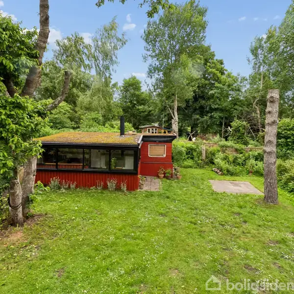 Et rødt træhus står stille i en frodig, grøn have med tæt bevoksning. Træer omgiver huset, og der er en lys blå himmel over den sceniske natur.