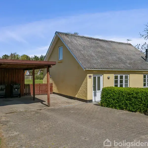 Gult hus med skråt tag står på en brolagt indkørsel ved siden af en carport. Haven er omkranset af grønne hække, og træer ses i baggrunden. Blå himmel ovenfor.