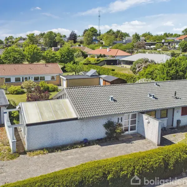 Hus med fladt tag og hvide murstensvægge, omgivet af grønne haver og nabo-bygninger i en forstadskvarter. En lille garage er ved siden af huset.