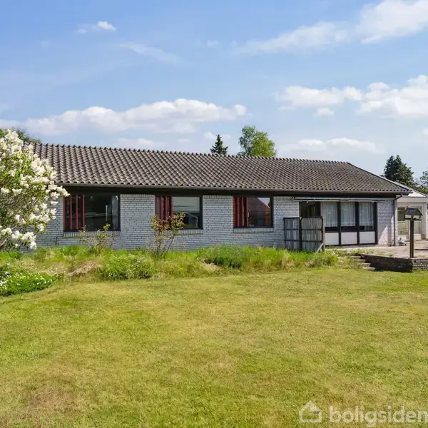 Et enkeltstående hus med hvide vægge og tegltag står i en have med en stor græsplæne. Til venstre står et blomstrende træ. Himlen er blå med spredte skyer.