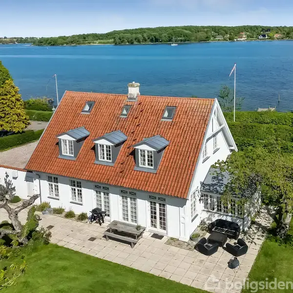 Et hvidt hus med rødt tag står ved en sø. Huset har flere vinduer på taget og terrasse med havemøbler. I baggrunden ses grønne træer og en blå sø.