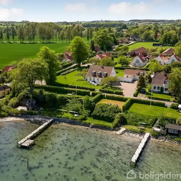 Kysthus står ved vandet med to bådebroer i en naturskøn forstad. Baggrunden har grønne marker og flere karakteristiske huse omgivet af træer og haver.