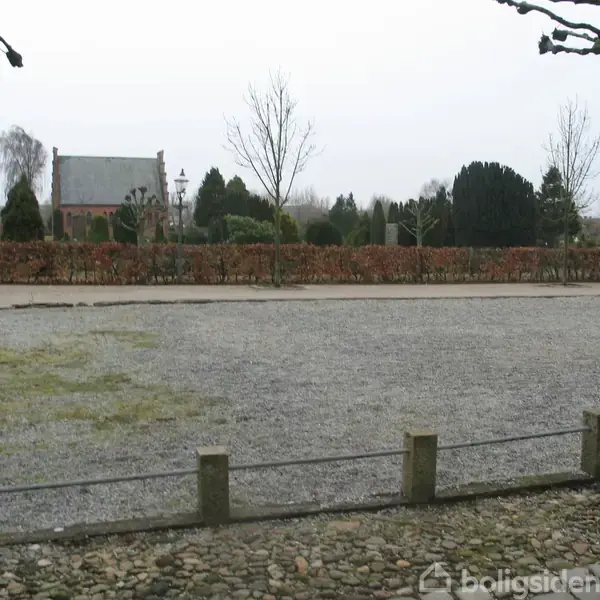 Grusplads, stille og ubenyttet, omgivet af hække og træer med en kirke i baggrunden. Mørkeblå bil delvist synlig til højre.