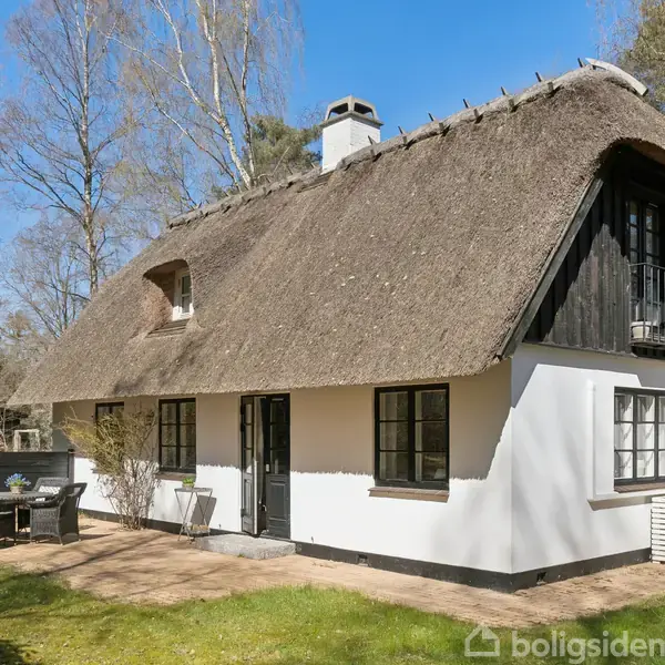 Hus med stråtag og hvid facade står i en skovklædt have. Ved siden af huset er der et bord med stole på en flisebelagt terrasse.