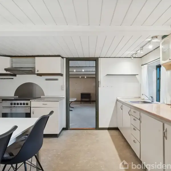 Køkken med hvidt træloft, sorte stole omkring et hvidt bord, moderne komfur og hvide skabe. En dør fører til en anden stue med mørkere indretning.