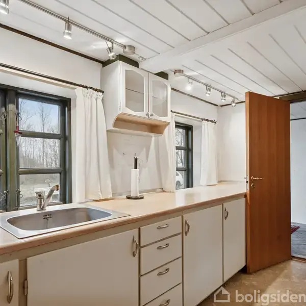 Et køkken med en vask og hvidt skabsmodul langs en væg; et åbent dør fører til en tilstødende stue. Vinduer med gardiner og udsigt udenfor.