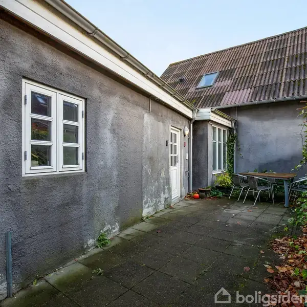 En gårdhave med grå bygning, flisebelagt terrasse og to vinduer. En hvid dør findes midt i billedet. Et havebord med stole står i det ene hjørne omgivet af grønt løv.