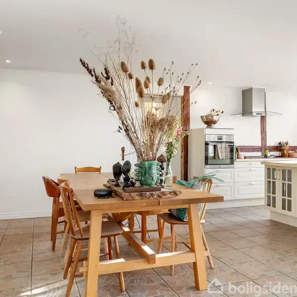 Et stort spisebord med tørrede blomsterarrangementer i centrum står i et lyst køkken-alrum med træmøbler og keramiske flisegulve. I baggrunden ses et komfur og køkkenø.