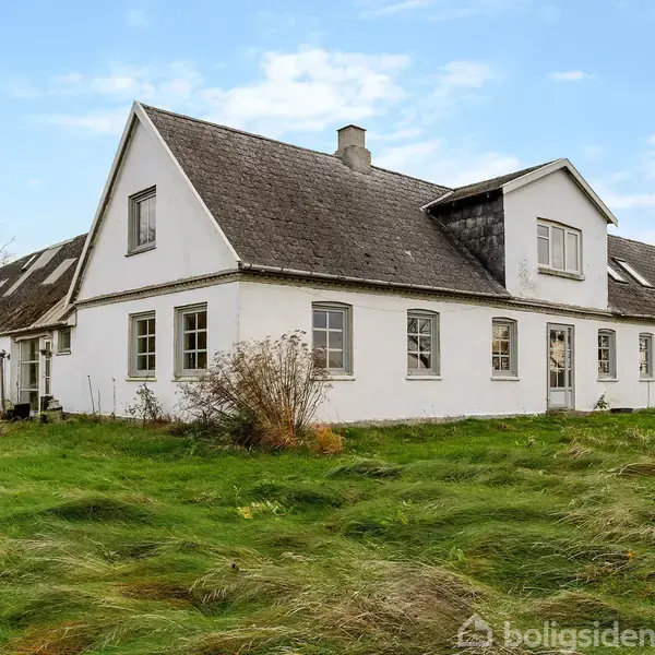 Hvidt, stort hus med skråt tag står stille i en grøn, overgroet have. Træer og en klar blå himmel danner baggrunden.