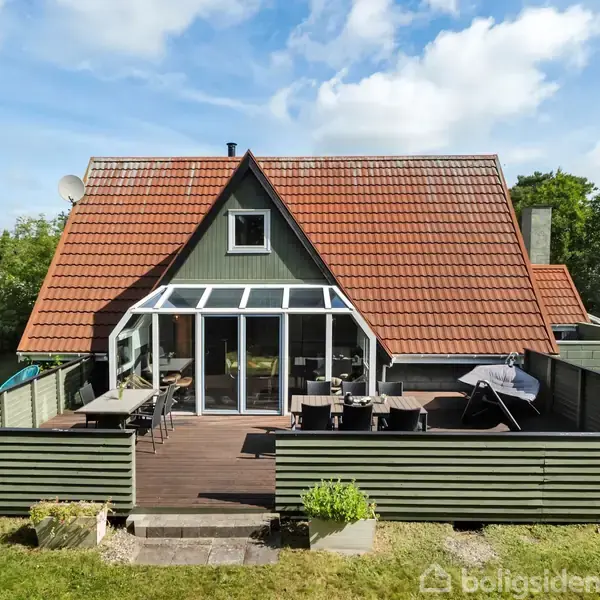 Hus med rødt tegltag og grøn facade, veranda med glasvægge, beliggende i en grøn have med træer i baggrunden. Terrassen har udemøbler og en grill.