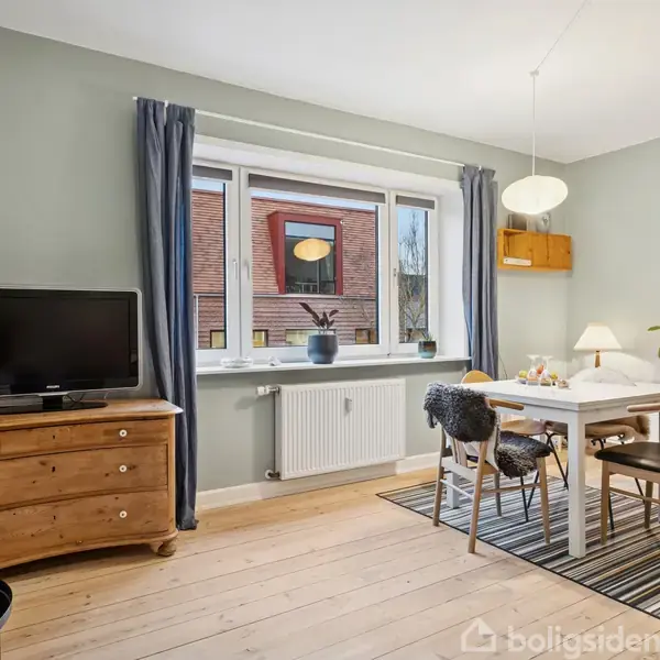 Fjernsyn placeret på en trækommode; ved siden af står et spisebord med stole, dækket med en dug, i en lys stue med vindue og grønne vægge.