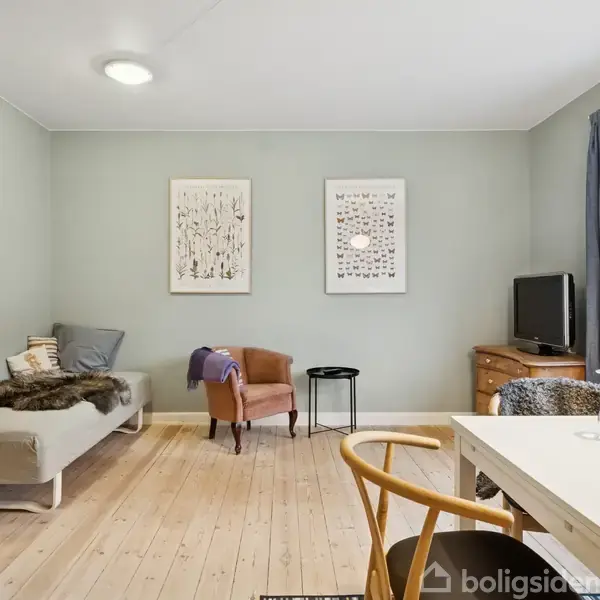 En stue med en sofa til venstre, en kommode og fjernsyn til højre, dekoreret med vægkunst. Bord, stole og et stort vindue danner et lyst og indbydende rum.