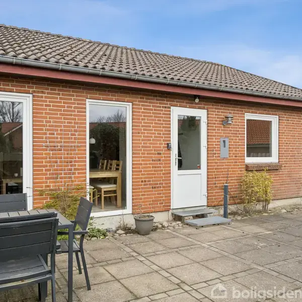 Et rødt murstenshus med et lavt, gråt tegltag. Foran huset er der en flisebelagt terrasse med et sort spisebordssæt. Store vinduer afslører et spisebord indenfor.