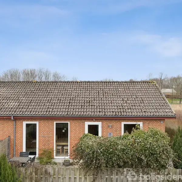 Rødt murstenshus har hæk og terrasse foran, beliggende i et åbent, landligt område med træer i baggrunden under en blå himmel.