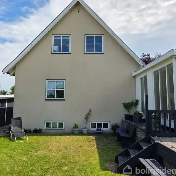 Hus med beige facade og grått tag ses fra baghaven, hvor en stol står på en græsplæne. Til højre findes en terrasse med blomsterkrukker. Det er solrigt med lette skyer.
