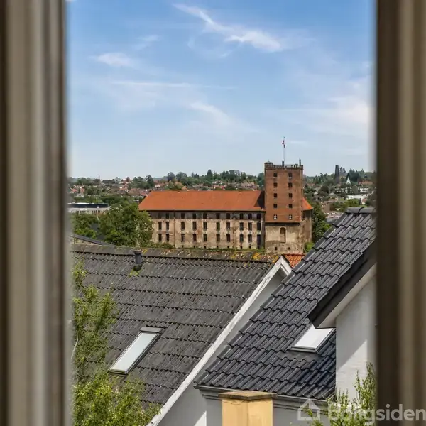 Et slot med rødt tegltag og tårn ses gennem et vindue. Slot er omgivet af huse med sorte tegltage, mens himlen er lyseblå med spredte skyer.