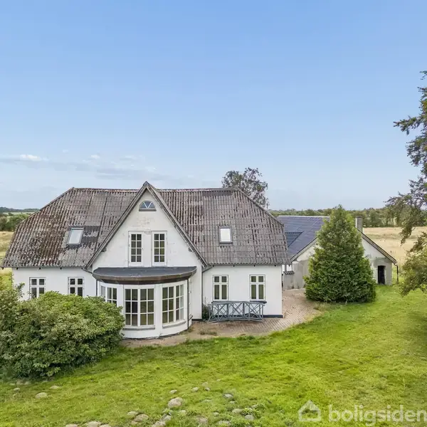 Stor, hvid villa med skråtag og karnap placeres i frodigt, landligt landskab. Der er en træterrasse og store græsarealer omkring huset. Solrigt vejr med blå himmel.