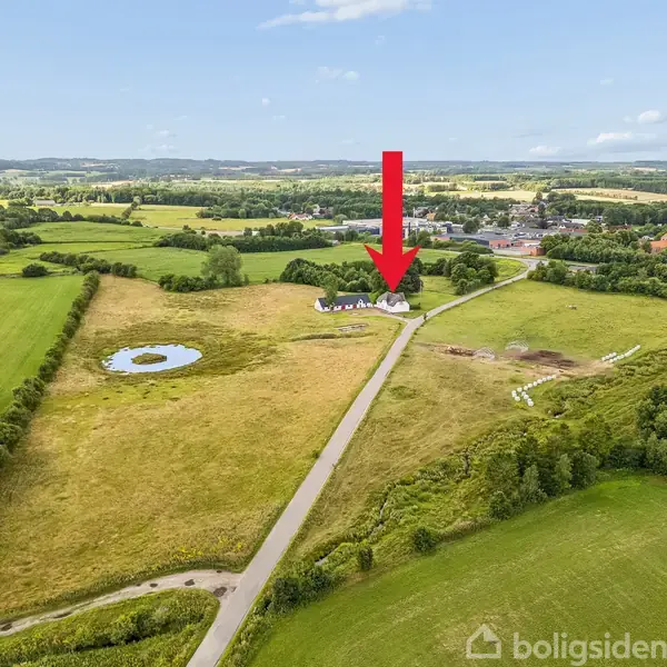 Hus markeret med rød pil langs en vej omgivet af grønne marker og et vandhul i landskabet. Landligt område med flere træer og bebyggelser i baggrunden under en klar himmel.