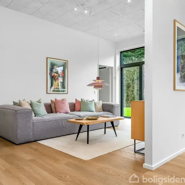 En grå sofa står i et lyst, moderne rum med træpaneler og farverige puder. Over sofaen hænger kunst, og ved sofaen står et sofabord på et tæppe nær en stor vinduesvæg.