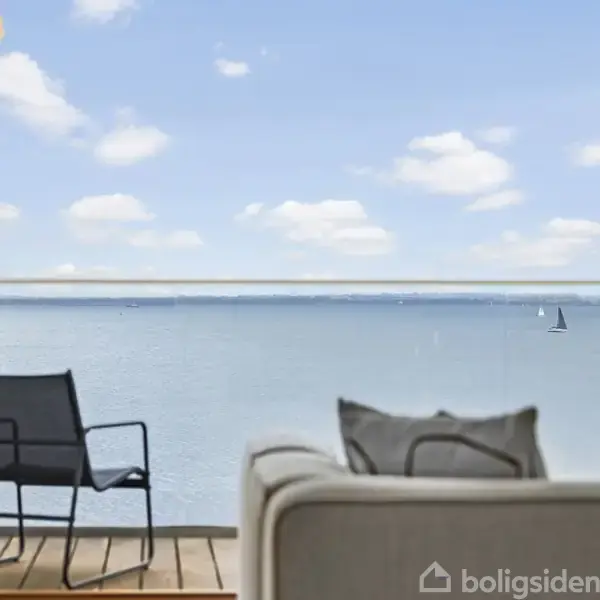Lænestol står stille med udsigt over havet; i baggrunden ses en sejlbåd på vandet under en skyfri blå himmel.